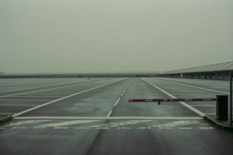 A wide, empty exhibition car park with painted lane markings and a raised barrier arm, hall roofs visible in the distance.